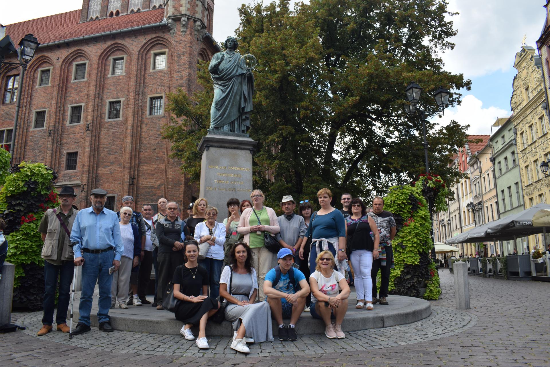Grupa uczestników wycieczki stoi przy pomniku na rynku przy pomniku Mikołaja Kopernika w Toruniu