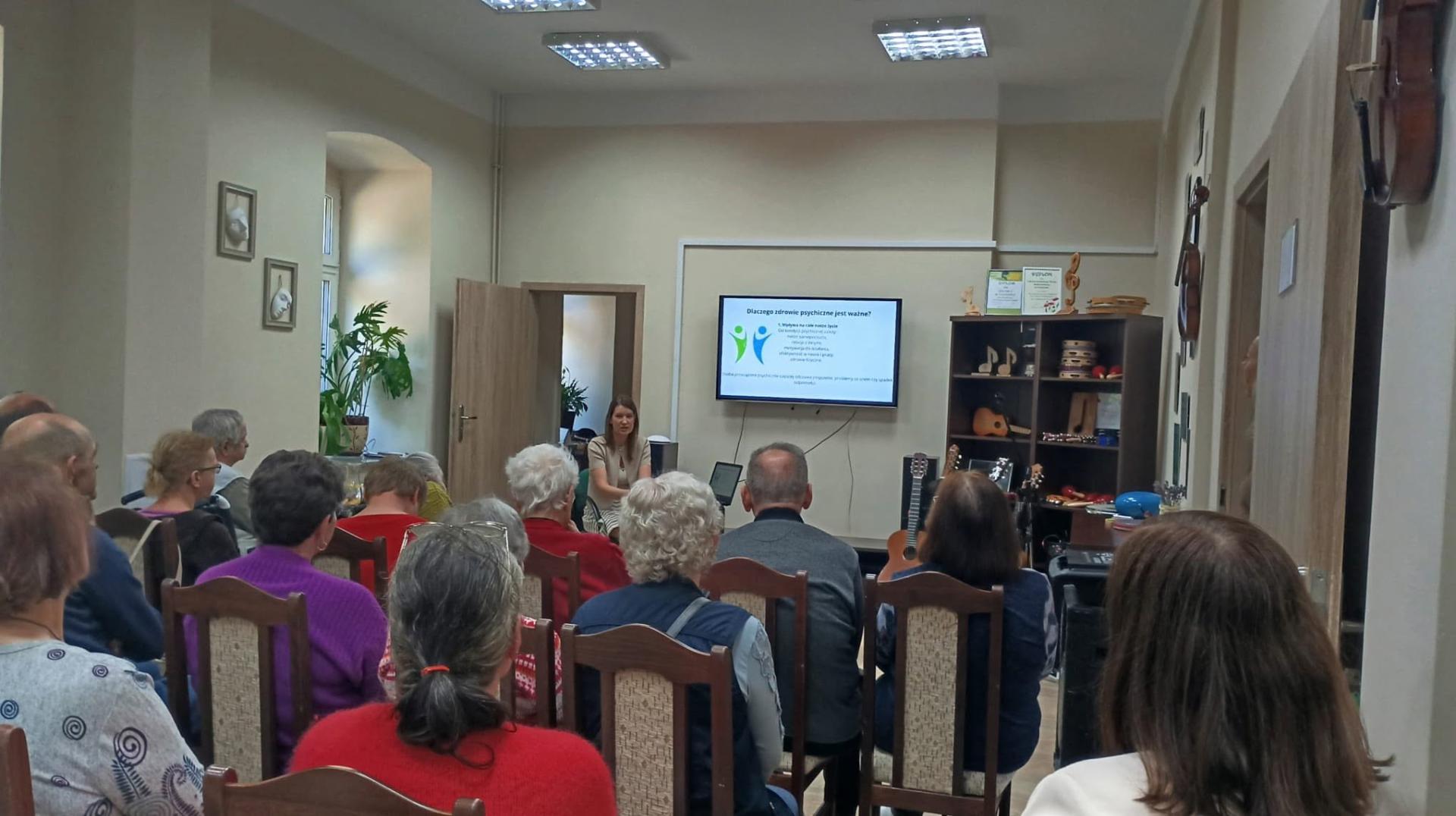 Na zdjęciu widać grupę osób siedzących w rzędach na krzesłach w sali i z uwagą słuchających wykładu na temat zdrowia psychicznego prowadzącego przez kobietę. Na przeciw siedzących słuchaczy widać wiszący na ścianie telewizor , na którym wyświetlony jest slajd z napisem -dlaczego zdrowie psychiczne jest ważne. Po lewej stronie siedzących osób widać parapet okna , na którym stoi doniczkowy kwiat, a na prawo od nich ,na ścianach wiszą skrzypce. Obok wiszącego telewizora stoi półka, na której znajdują sie instrumenty muzyczne .