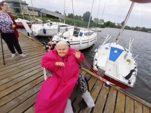 Niepełnosprawna kobieta na wózku , w różowym płaszczu przeciwdeszczowym macha do zdjęciu, w tle widać jezioro i białe łódki.