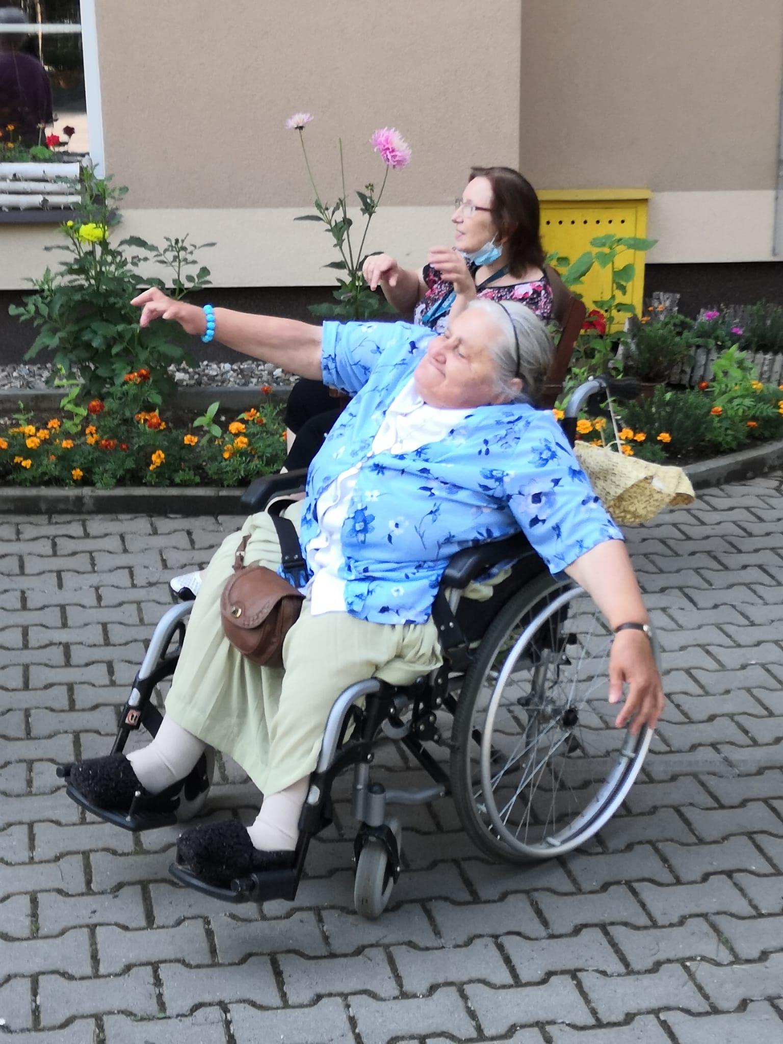 Kobieta siedząca na wózku inwalidzkim powtarza ruchy taneczne wykonujące przez instruktora tańca. Po jej prawej stronie siedzi kobieta i robi to samo .