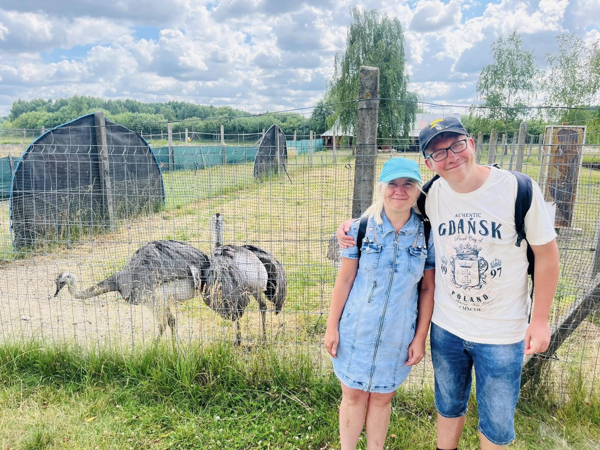 Młoda, uśmiechnięta  kobieta ubrana w krótką , dżinsową sukienkę stoi obok młodego , uśmiechniętego mężczyzny . Obydwoje stoją na trawie , na głowie mają czapki z daszkiem , a na plecach plecaki. Za nimi widać dwa chodzące na wybiegu strusie.