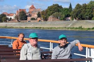 Trzech mężczyzn siedzi na statku . W tle widać panoramę Torunia.