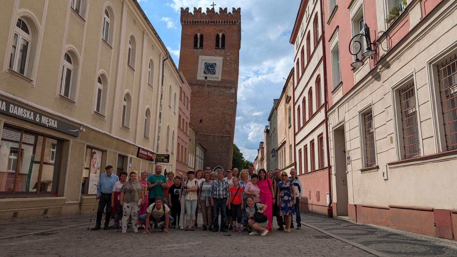 Na zdjęciu widać uśmiechniętą grupę kobiet i mężczyzn , uczestników projektu pozujących do zdjęcia . Ubrani są w cienkie, letnie ,kolorowe ubrania . Stoją na ulicy pomiędzy budynkami mieszkalnymi . Za nimi widać krzywą wieże.