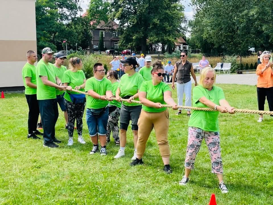 Słoneczny dzień. Grupa kobiet i mężczyzn, drużyna sportowa ,uczestnicząca w projekcie stoi kolejno jeden za drugim . Ubrani są w zielone sportowe koszulki z krótkim rękawkiem oraz zielone czapki. Każda osoba w ręku trzyma linę do przeciągania .  Stoją na zielonej trawie , z tyłu za nimi widać białe ławki oraz zielone drzewa.