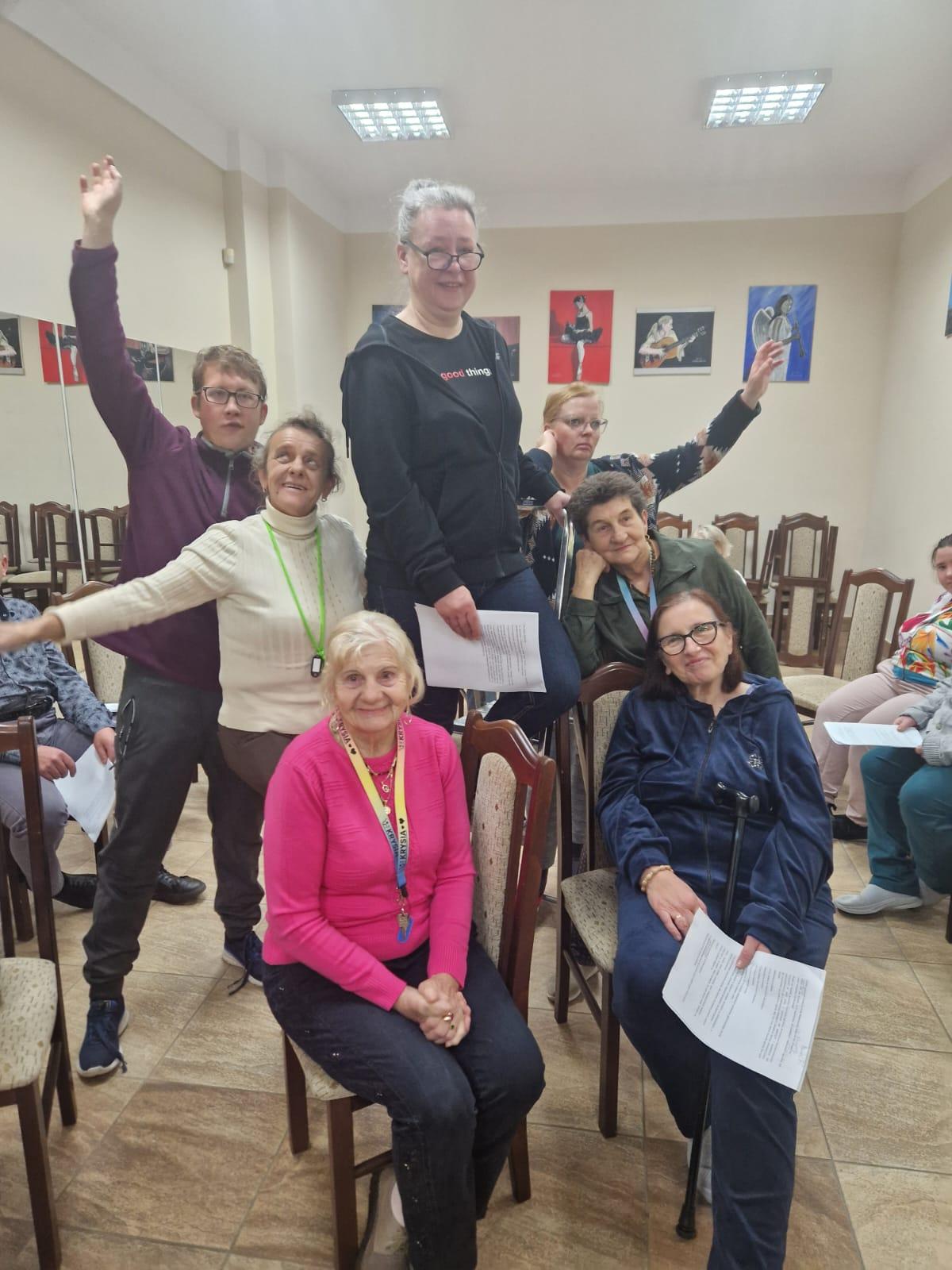 Na zdjęciu widać pozujących uczestników projektu . Na pierwszym planie , na krzesłach siedzą dwie , uśmiechające się kobiety . Jedna z nich, siedząca po lewej stronie , blondynka, ubrana jest w różowy sweter i czarne spodnie . Na szyj na zawieszone klucze . Druga z kobiet , brunetka w okularach ma na sobie granatowy dres. W ręku trzyma kartkę papieru. Za nią widać zadumaną kobietę , podpierającą ręką brodę. Obok niej stoi kobieta , która uśmiechając się stoi na krześle, a w ręku trzyma kartkę papieru> Stojąca obok niej, również uśmiechnięta kobieta w białym golfie spogląda do góry , a mężczyzna , ma uniesioną prawą rękę do góry . Za uczestnikami projektu widać stojące krzesła oraz ścianę , na której wiszą kolorowe obrazy .