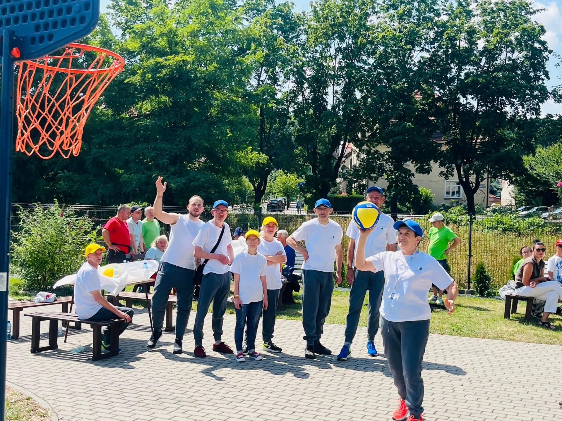 Piękny , słoneczny dzień. Drużyna sportowa, grupa kobiet i mężczyzn  ubranych w szare spodnie dresowe, białe sportowe koszulki z krótkim rękawem oraz żółte i zielone czapeczki z daszkiem stoją kibicując koleżance z drużyny, która stoi z uniesioną prawą ręką, w której trzyma piłkę . Próbuje nią trafić do stojącego przed nią kosza do piłki. Za drużyną widać zielone drzewa oraz siedzącą na ławkach widownię .