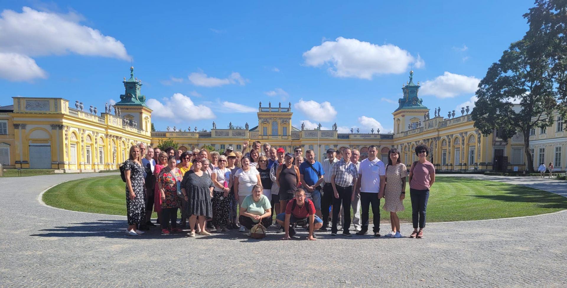 Pogodny, słoneczny dzień . Uśmiechnięta grupa kobiet i mężczyzn , stoją obok siebie i pozują do zdjęcia . Z tyłu za nimi widać okrągły, zielony trawnik oraz Muzeum Pałacu Króla Jana III w Wilanowie.