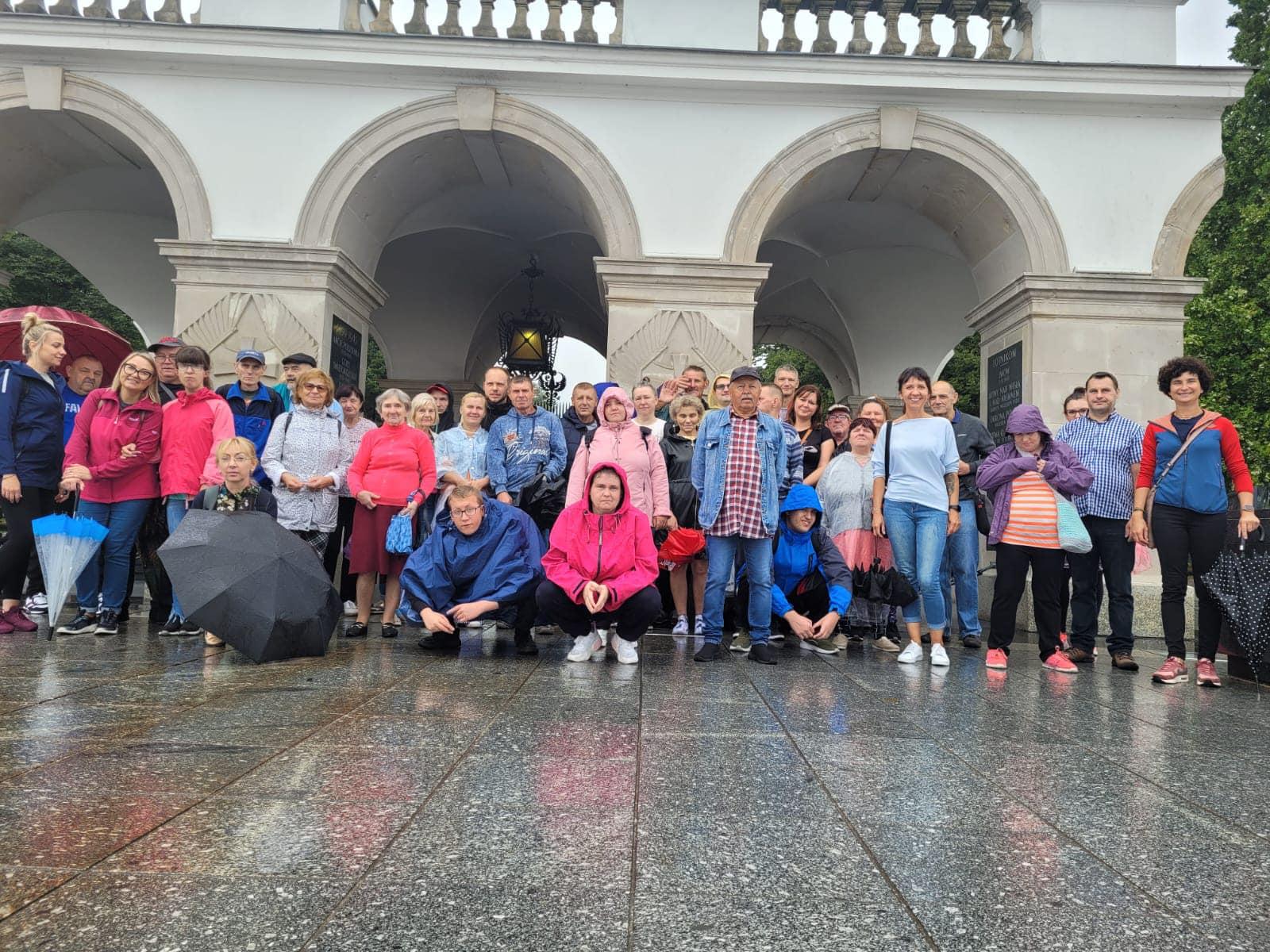 Deszczowa pogoda. Grupa kobiet i mężczyzn stoją obok siebie i pozują do zdjęcia . Ubrani są w kolorowe bluzy i płaszcze przeciwdeszczowe. Kilka osób trzyma w ręku parasolkę. Za nimi widać Grób Nieznanego Żołnierza.