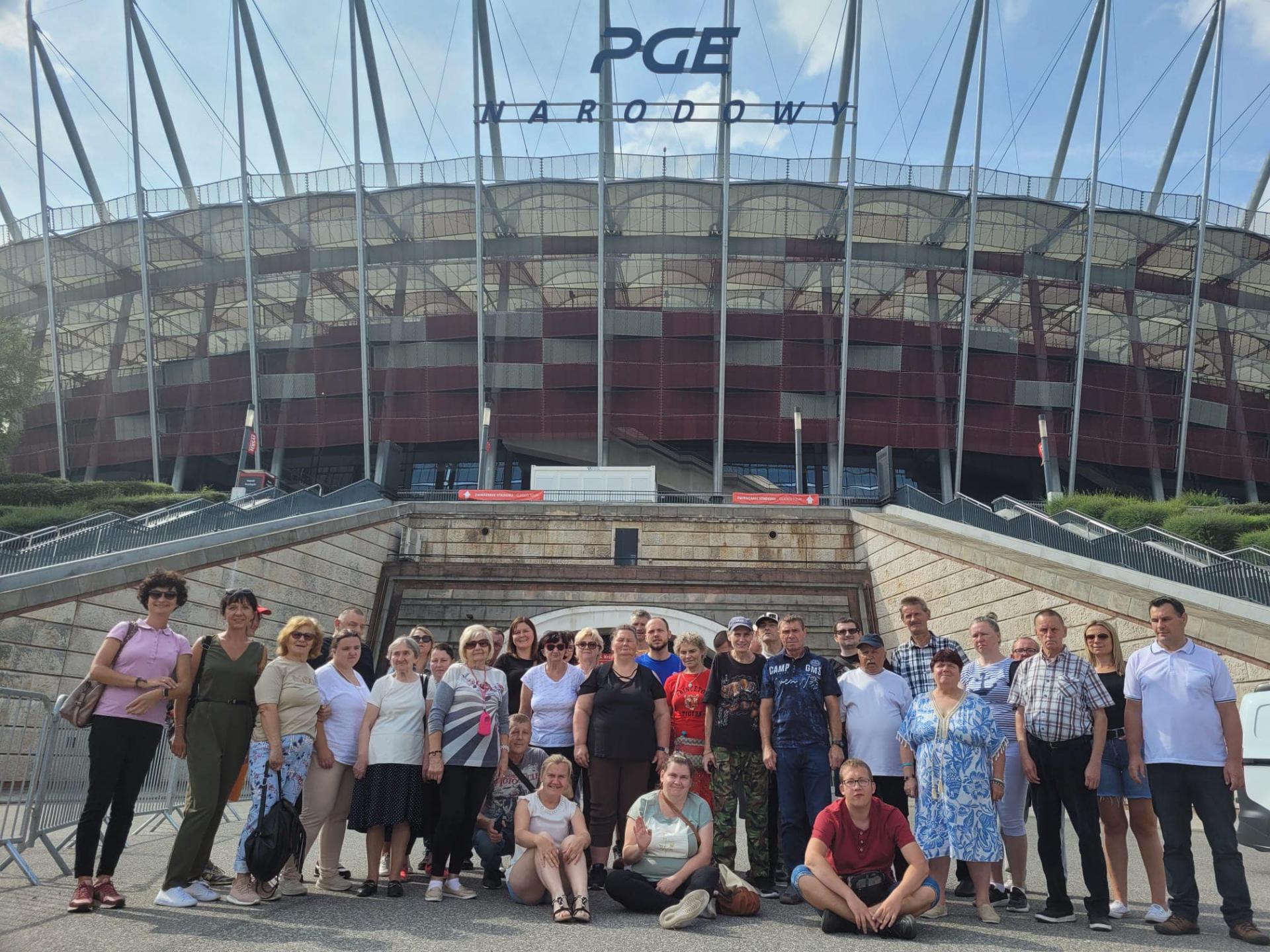 Pogodny dzień . Uśmiechnięta grupa kobiet i mężczyzn , stoją obok siebie i pozują do zdjęcia . Z tyłu za nimi widać biało-czerwony  Stadion PGE Narodowy.