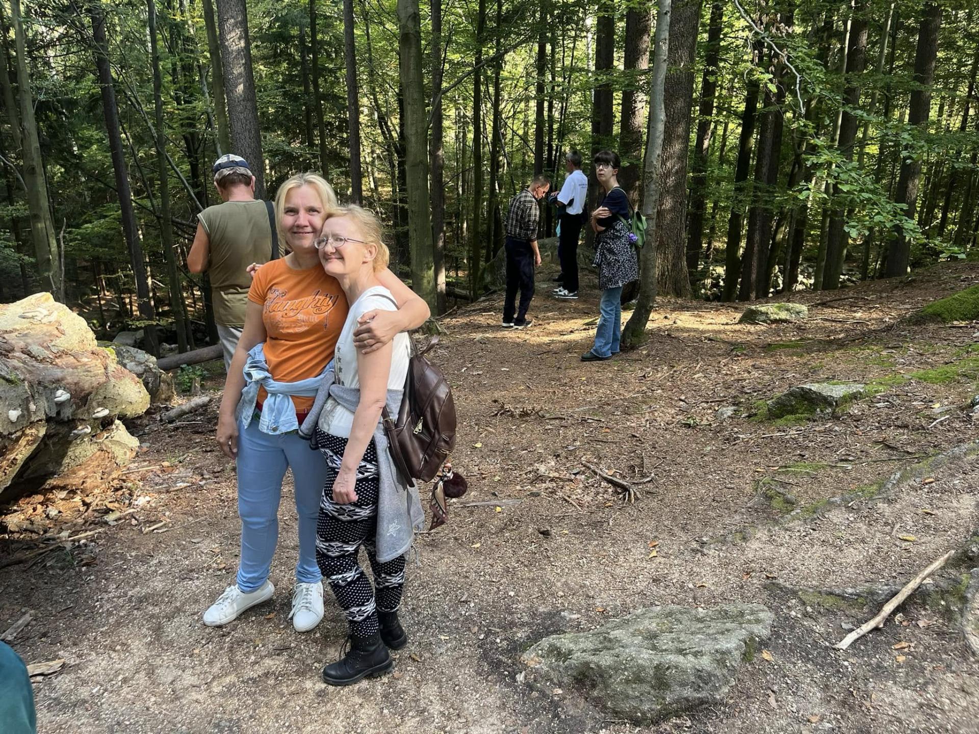 Dwie kobiety stoją wśród drzew pozując do zdjęcia . Jedna obejmuje z uśmiechem drugą . Za nimi widać kobietę z założonymi rękoma , opierającą się o drzewo, a obok mężczyznę odwróconego plecami . Przez drzewa widać przebijające się promienie słońca.