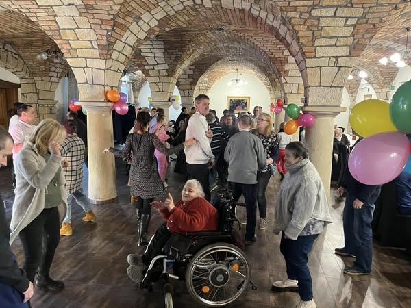 Na sali bankietowej widać kolorowo ubrane , tańczące osoby . Na ceglanych kolumnach zawieszone są kolorowe balony. Po środku tańczących osób widać kobietę siedzącą na wózku inwalidzkim poruszającą rękoma w rytm muzyki.