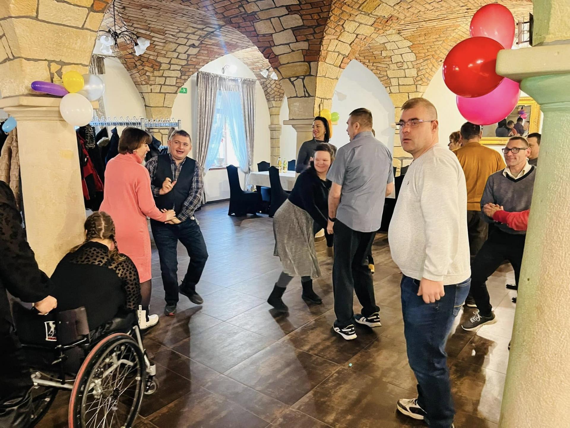 Na sali bankietowej widać kolorowo ubrane , tańczące osoby, również tańczące pary . Na ceglanych kolumnach zawieszone są kolorowe balony. Po  lewej stronie  widać kobietę siedzącą na wózku inwalidzkim. Wózek trzymany jest z tyłu przez inną kobietę .