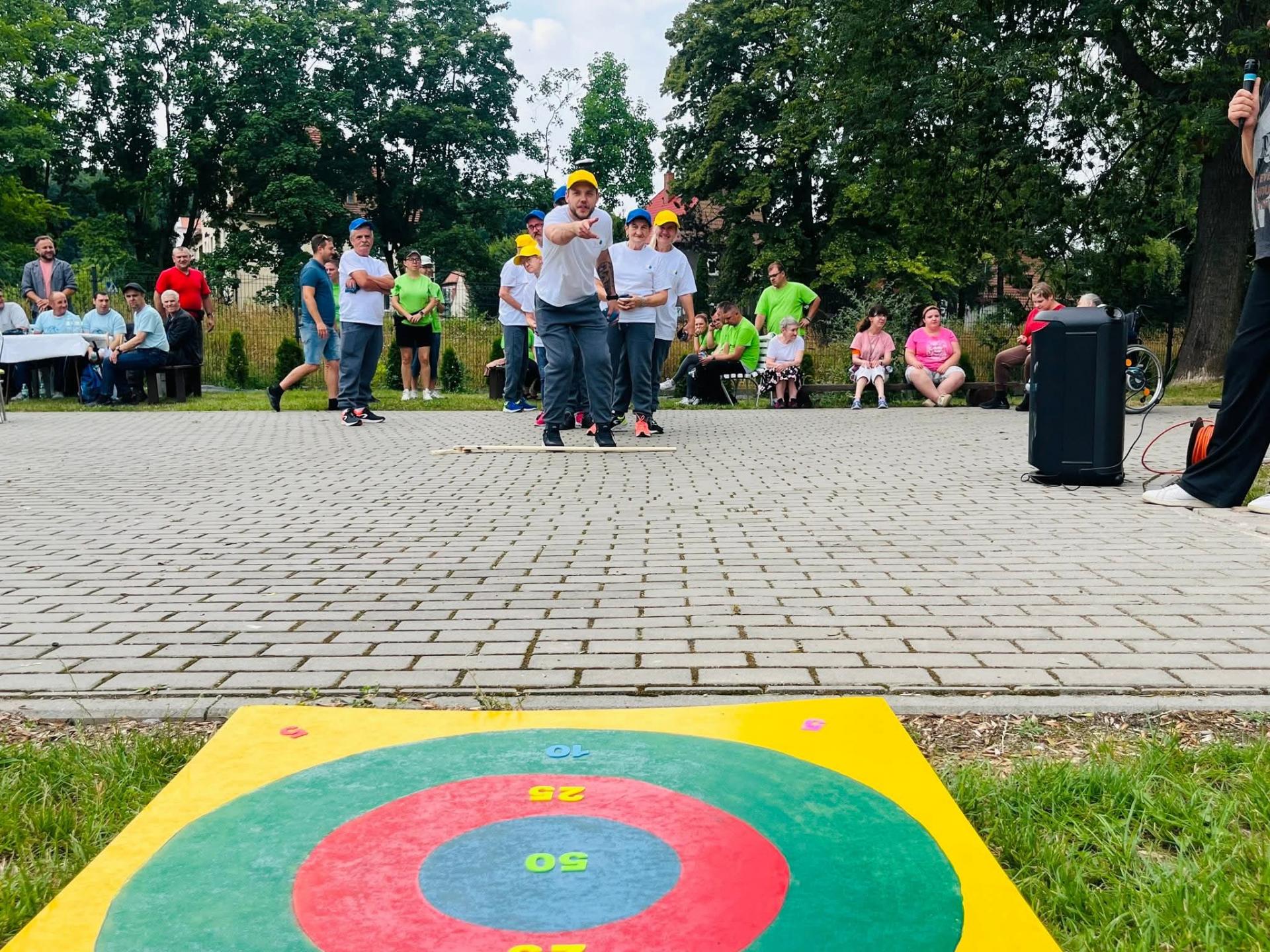 na pierwszym planie widać leżącą na ziemi , okrągłą , kolorowa tablicę punktową. W niedużej odległości od niej stoi nachylony do rzutu mężczyzna.  Ubrany jest w szare, dresowe spodnie , białą sportową koszulkę . Na głowie ma żółtą czapeczkę z daszkiem apeczkę z daszkiem . Za nim widać kibicującą mu drużynę sportową . W tle widać siedzącą na ławkach publiczność i inne drużyny.